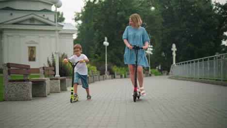 a happy mother and her son ride scooters side by side on a paved walkway, a younger boy on a bicycle follows nearby