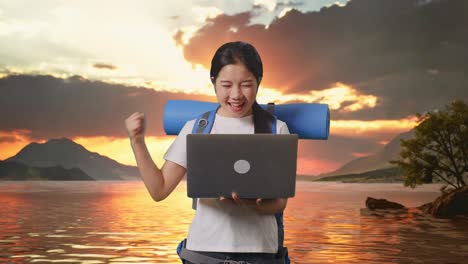 excursionista asiática con mochila de montañismo mirando una computadora portátil y gritando meta celebrando el éxito en un lago