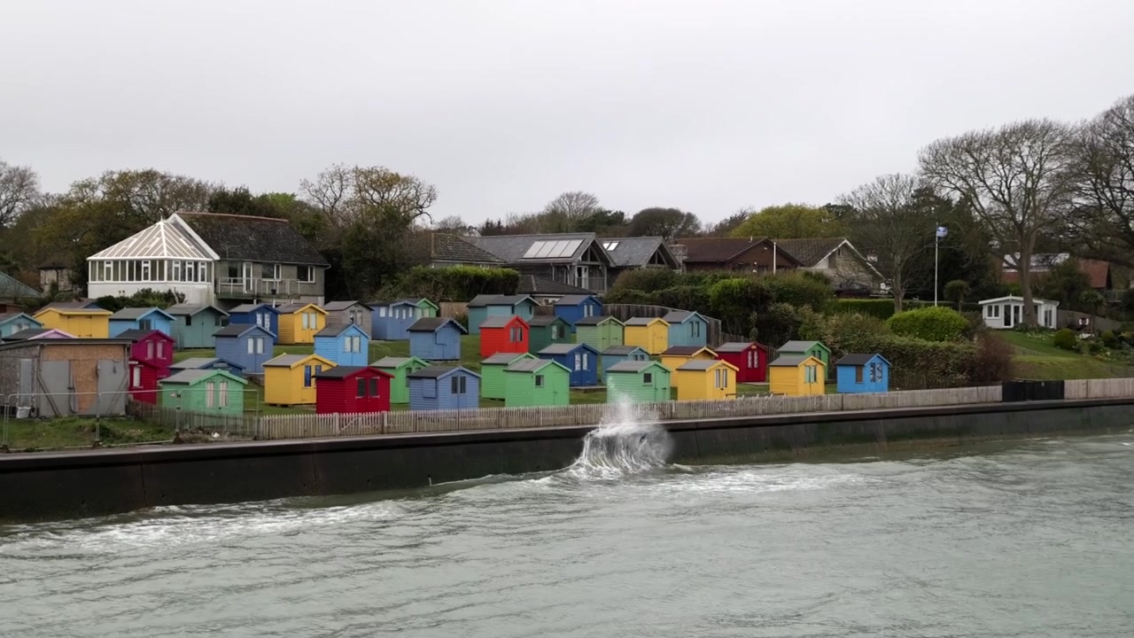 Colorful beach huts along Isle of Wight waterfront with waves splashing under cloudy sky.