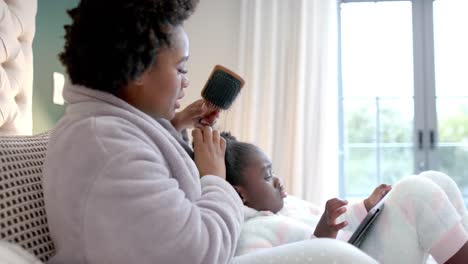 Happy-african-american-mother-combing-hair-of-daughter-using-tablet-in-bedroom,-slow-motion