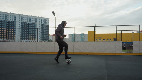 athlete training with football on sport arena tapping ball forward with precision, showcasing control and agility, background features urban buildings, yellow wall, people walking, and fencing