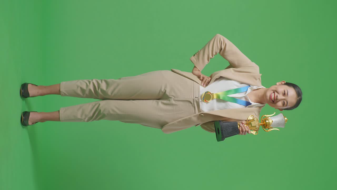 금메달과 트로피를 입은 슈트를 입은 아시아 사업가 여성이 한 팔로 카메라를 향해 미소 짓고 있으며, 아보는 스튜디오의 녹색 스크린 배경에서 첫 번째 우승자로 포즈를 취하고 있습니다.