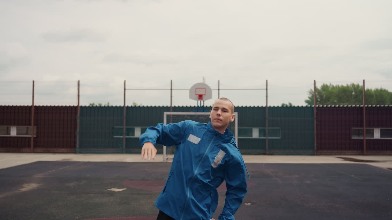 Caucasian footballer on wet court practicing juggling and stretches under cloudy sky, blue rain jacket, empty playground, chainlink fence, focused expression, cinematic moody light, subtle drizzle