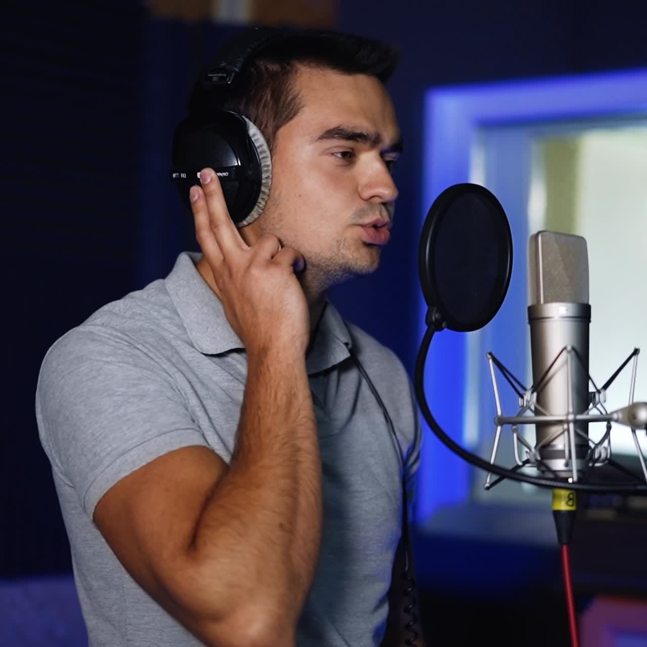 Caucasian man in grey shirt sings to the professional microphone. Singer performs a song holding by the headphones on his head