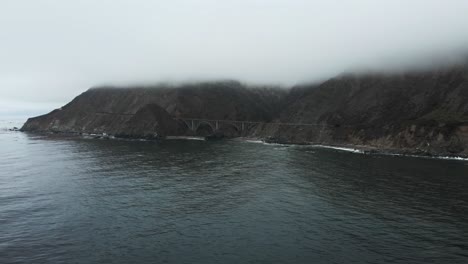 A drone shot strafing and entering a low cloud ceiling revealing the Bir Sur coastline with turquoise Pacific ocean saltwater with waves crashing against cliffs off the historic Highway 1