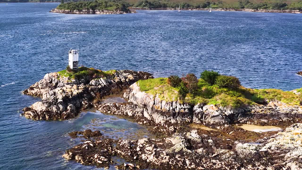 Drone circles rocky island with lighthouse, clear daylight, blue sea, vibrant greenery, smooth movement