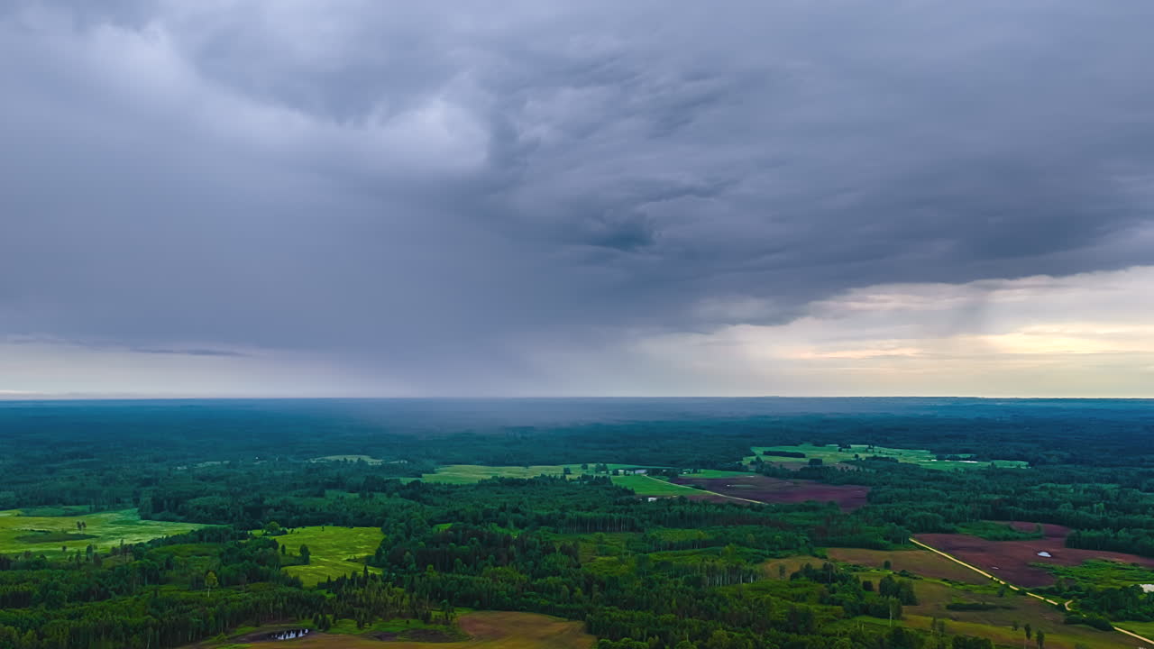 An aerial timelapse captures dramatic grey storm clouds moving quickly over a vast Latvian landscape of dense green forests and agricultural fields, signaling impending rain