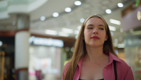 lady in pink dress slightly raising her hand while holding shopping bags, walking confidently with a subtle smile in a bustling mall, blurred background with people and store lights in view