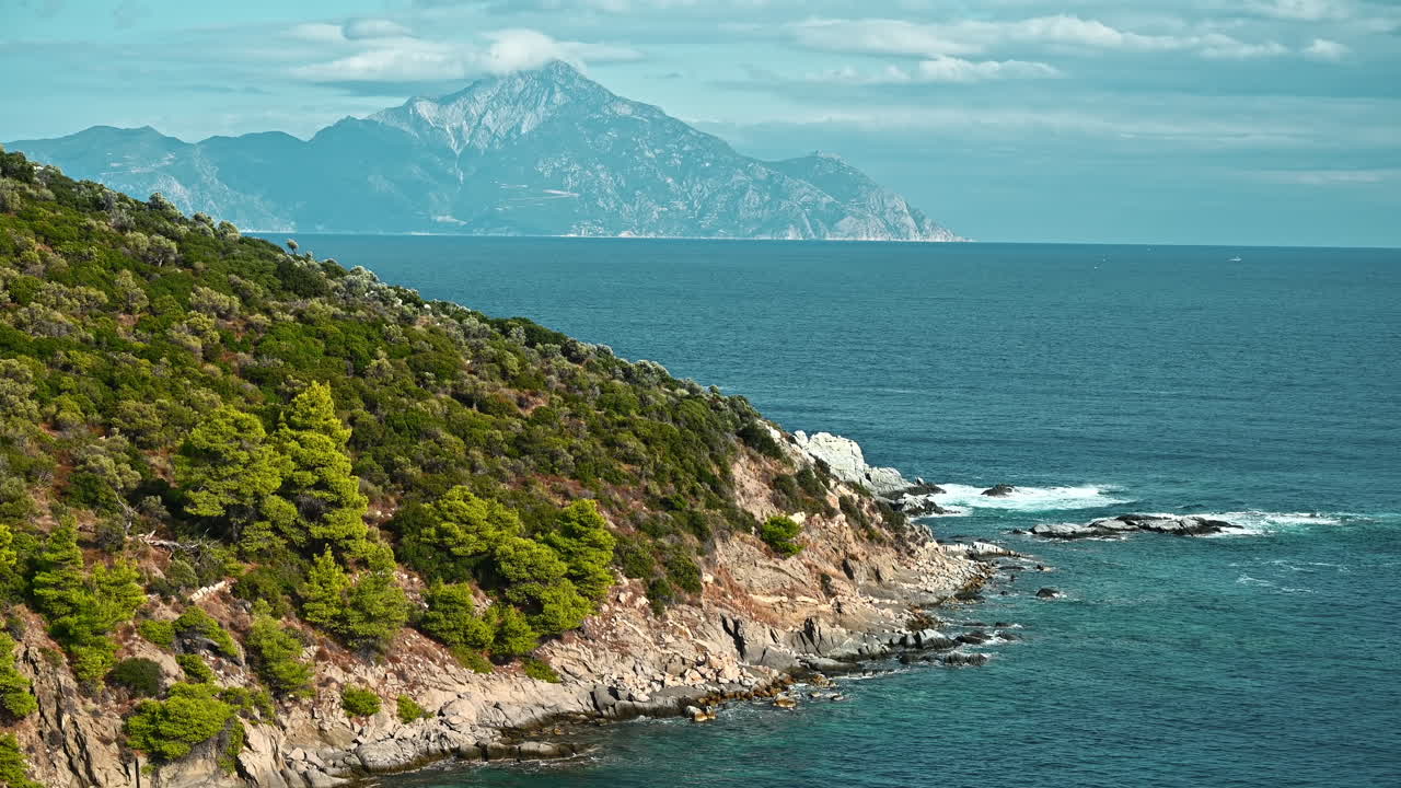 Aegean sea rocky coast with greenery around, bushes and trees, blue water with waves, mountain on the background, Greece