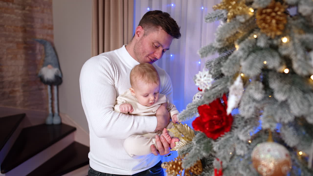 Caucasian man wearing warm white sweater holding a baby in light clothes. Loving daddy waves his barefoot son, kisses him on the head and shows the beautifully decorated Christmas tree. Top view.
