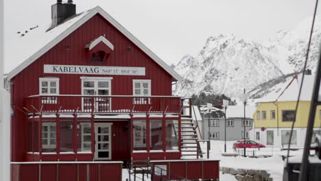 Icy-landscape-of-the-village-of-Kabelvåg,-Norway-with-a-red-house-in-the-foreground-and-snow-capped-mountains-in-the-background