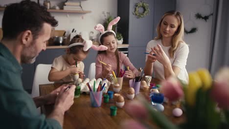 familia caucásica de cuatro personas charlando y decorando huevos de pascua en la cocina doméstica.