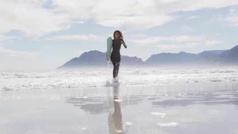 mixed race woman running from the sea onto beach carrying surfboard