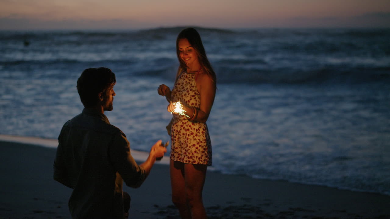 새해 전야를 축하하며 스마트폰을 사용하여 춤추는 폭죽을 들고 있는 여자친구의 사진을 찍는 청년은 일몰 슬로우 모션으로 해변에서 함께 장난스럽게 웃고 있습니다