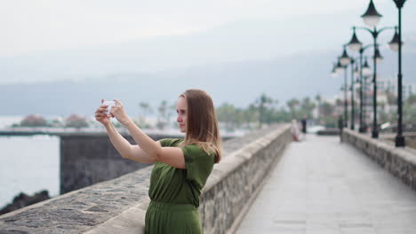 Ein-Schönes-Mädchen-In-Einem-Grünen-Kleid-Dreht-Ein-Video-Für-Einen-Blog-Auf-Einem-Mobiltelefon,-Steht-Am-Wasser-In-Der-Nähe-Des-Ozeans-Und-Lächelt