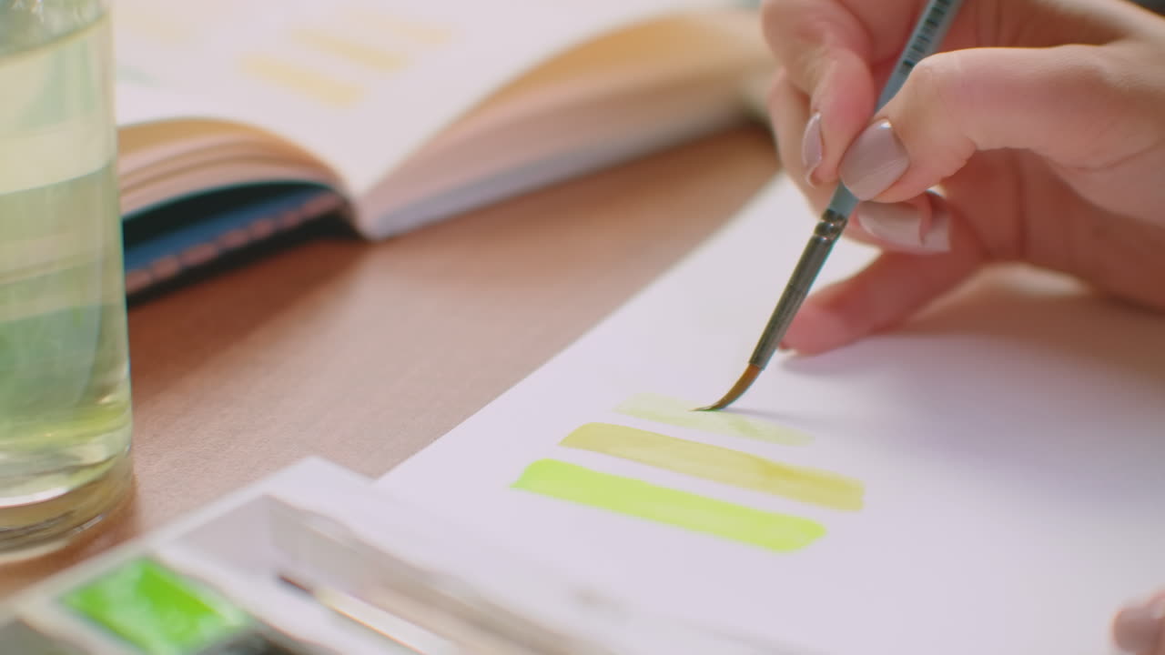 브러쉬와 페인트를 가진 젊은 여성은 로고 디자인 또는 벽에 적합한 색조를 선택합니다. 미래의 디자인을위한 색상과 색조의 선택. 큰 창문이있는 사무실의 여성 컬러리스트는 브러쉬를 사용하여 노트북에 그림을 그립니다.