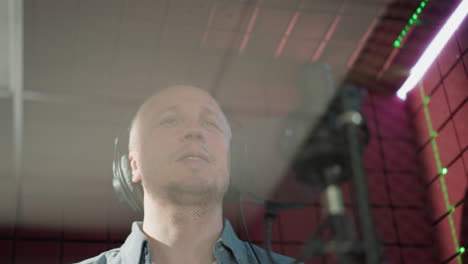 a man in a recording studio wearing a blue long sleeve shirt and black headphones, singing passionately in front of a microphone with a red acoustic panel background, under soft lighting