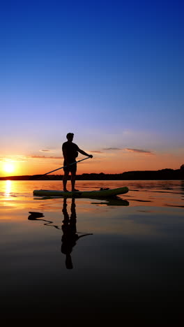 Man standing on sup board reflects in the waterscape at sunset. Paddle boarding practice in summer. Low angle view. Vertical video.