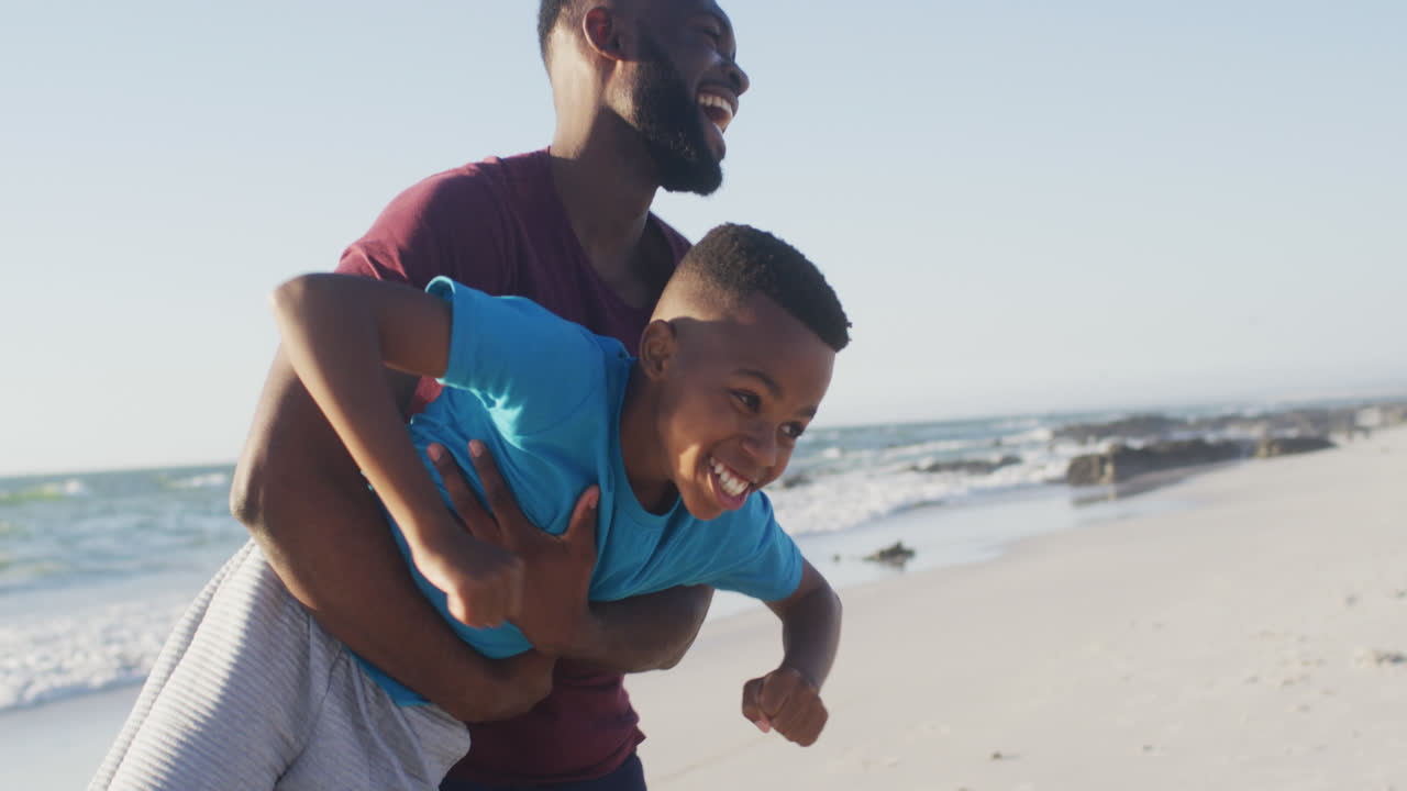 Video of happy african american father and son having fun on beach