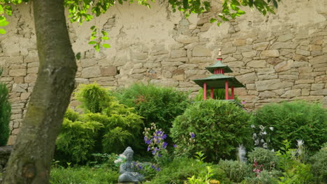 Ornamental-Chinese-pergoda-and-buddha-statue-among-garden-shrubs-and-flowers