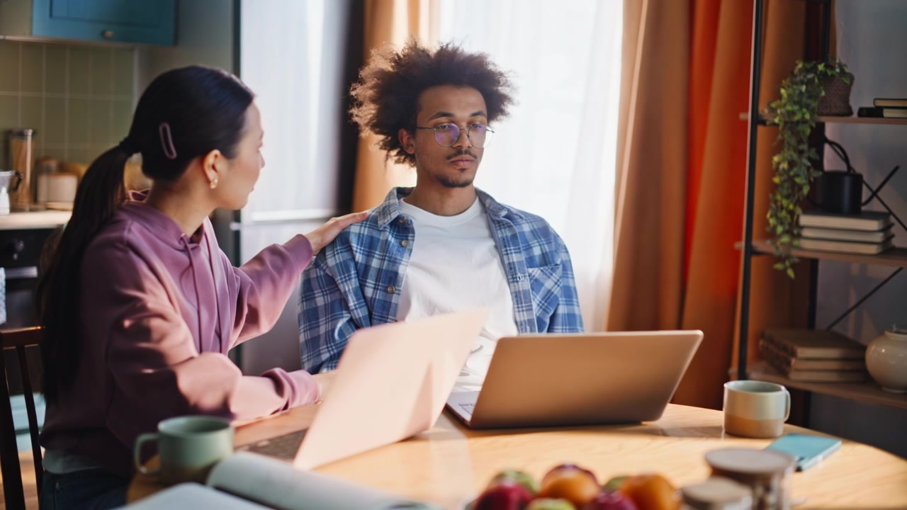Multiethnic couple working laptops home. Man frustrated by bad news online.