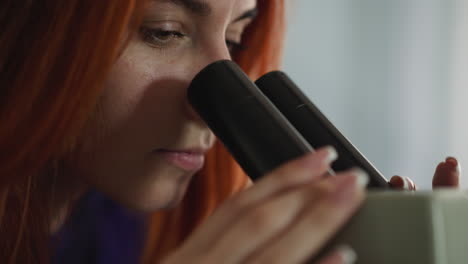 mujer enfocada con cabello rojo mira a través del microscopio en el laboratorio