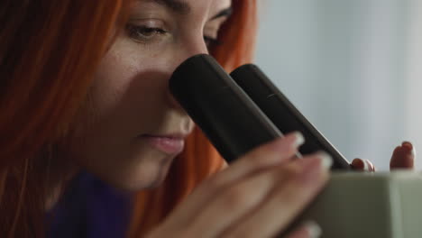 focused woman with red hair looks through microscope in lab