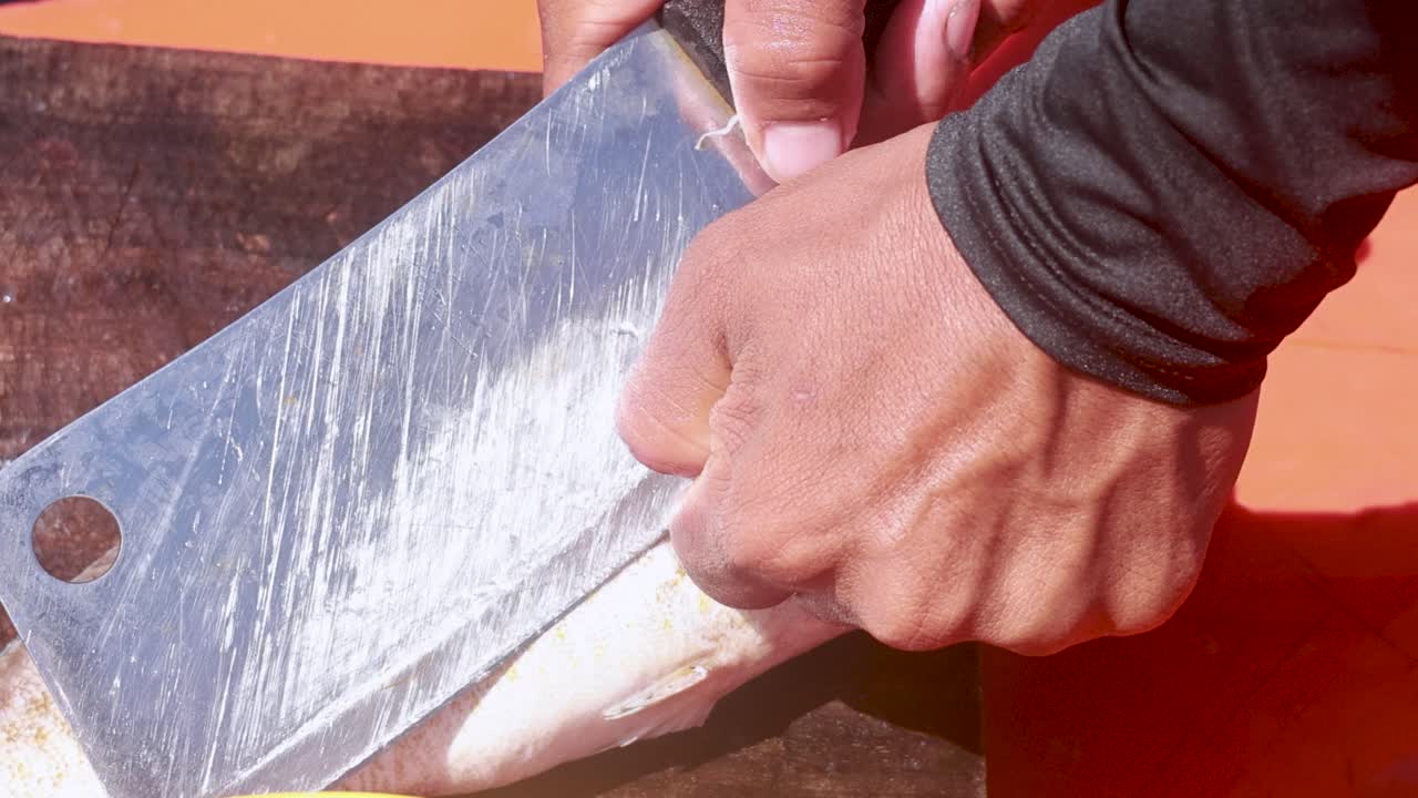 A person skillfully cleans a fish using a cleaver on a wooden surface in bright daylight