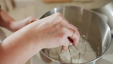 close up of hands dipping metal mixer attachment into bowl with thick mixed flour and lifting it out, coated with creamy mixture, showcasing baking preparation in process