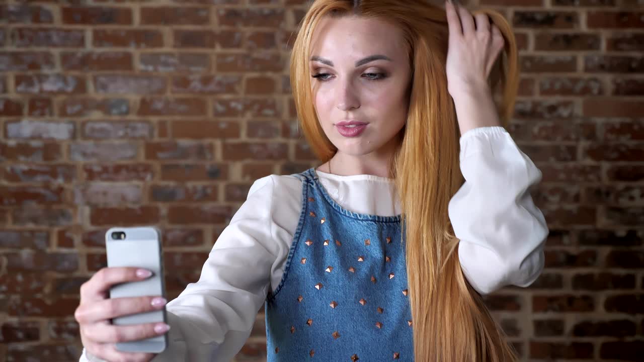 joven chica rubia dulce está haciendo selfie en el teléfono inteligente, fondo de ladrillo, concepto de comunicación