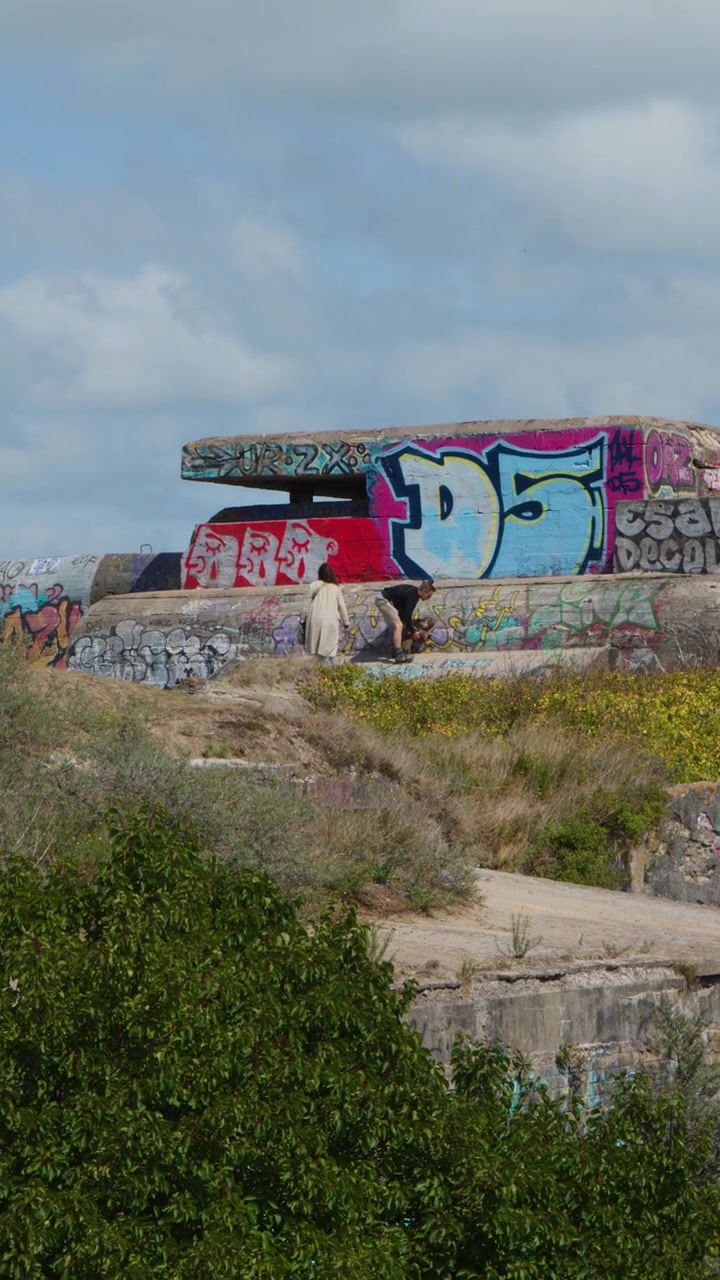 Individual with dog walks past colorful graffiti on abandoned concrete bunker, overgrown coastal landscape, daylight