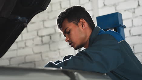 automobile engineer in blue uniform leans over car engine while conversing in workshop, focused mechanic looks engaged in discussion with bright light shining in background