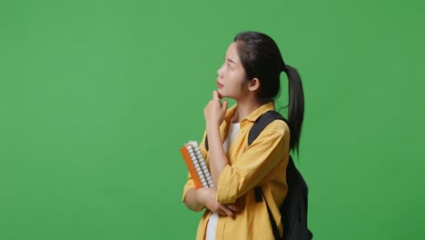 vista lateral de una estudiante asiática con una mochila y algunos libros pensando y mirando a su alrededor mientras está de pie en el estudio de fondo de la pantalla verde