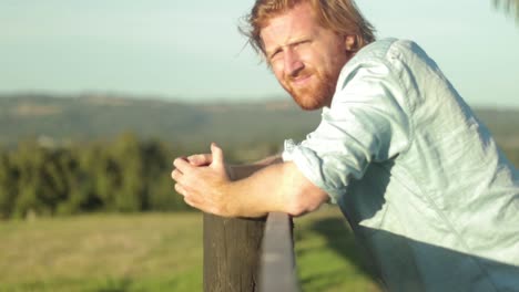 Un-Joven-Agricultor-Con-Una-Camisa-Azul-Descansa-Sobre-Un-Poste-De-Madera-Y-Mira-A-Través-Del-País-En-Cámara-Lenta