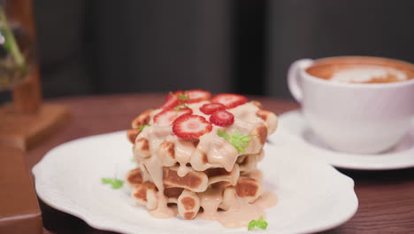 stack of creamy waffles topped with strawberry slices and garnished with herbs placed beside white cup of latte on brown wooden table near brown book in elegant indoor setting