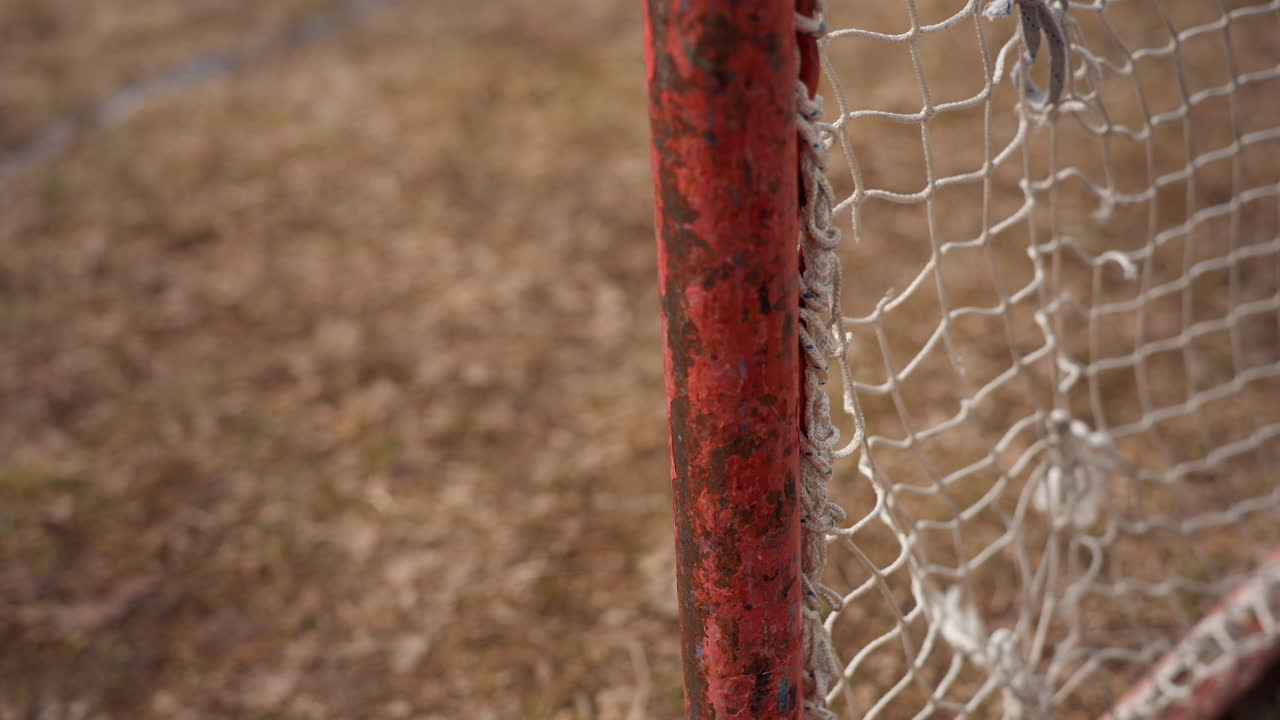 worn red goal on field, aged scarlet goal frame with frayed white netting on rustic terrain, weathered maroon goalpost featuring tattered netting amidst textured urban playing surface