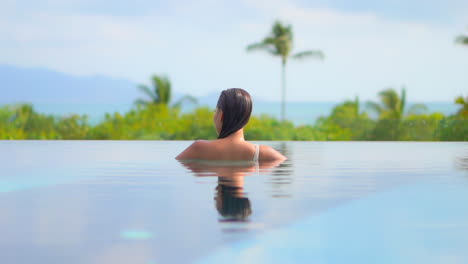 Una-Mujer-Irreconocible-Dentro-De-Una-Piscina-Infinita-Mirando-La-Asombrosa-Vegetación-Tropical-En-El-Fondo-De-Las-Colinas-Durante-El-Día,-Reflejo-Corporal-En-La-Superficie-Del-Agua