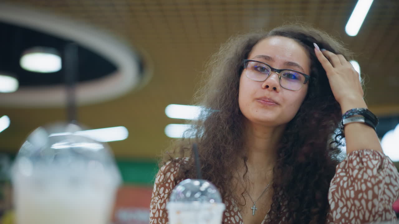 mujer beba una bebida cremosa con paja, ajustando su cabello y sonriendo en un centro comercial animado, creando un ambiente relajado y agradable con otra bebida cerca, fondo borroso y luces bokeh suaves