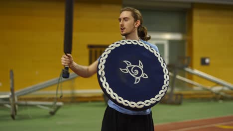 young man with long hair practices larp foam combat blue sword and blue shield inside a gym while the camera pans right