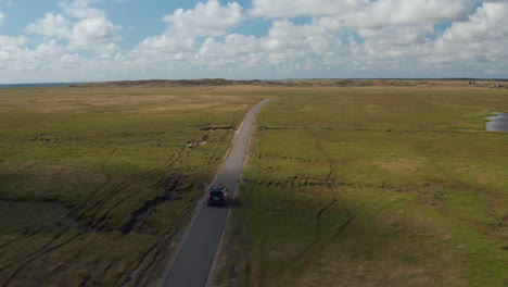 Aerial-footage-of-car-going-on-narrow-pathway-leading-in-flat-grasslands.-Green-pasture-in-coastal-landscape.-Denmark