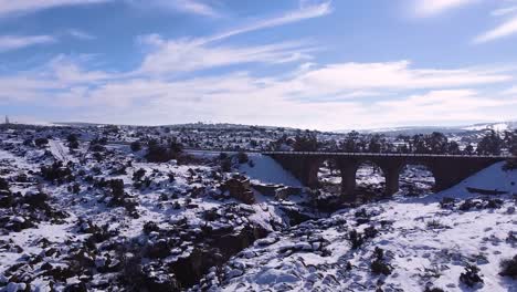 Aerial-view-of-a-drone-flying-over-a-beautiful-sunny-snowy-field-with-a-river,-an-old-bridge-and-an-old-house