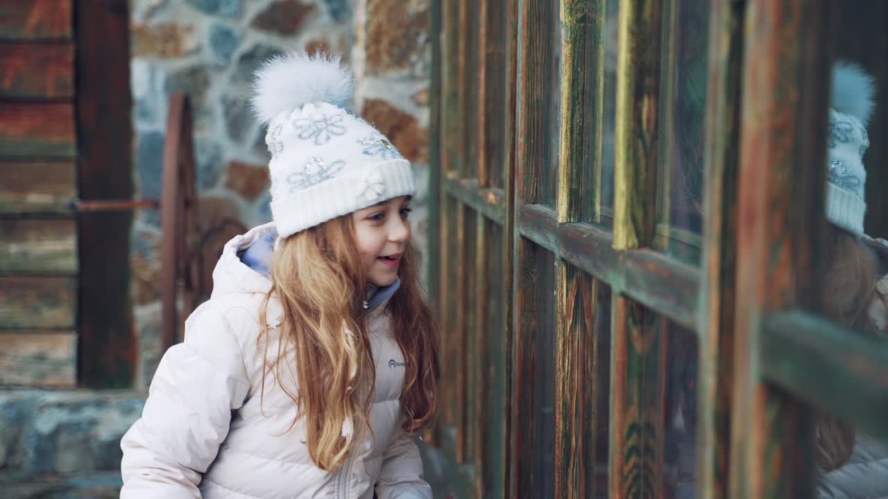 A little girl with an envelope in her hand is peeping through the window to elves at day before the New Year in the street on the background of snow.