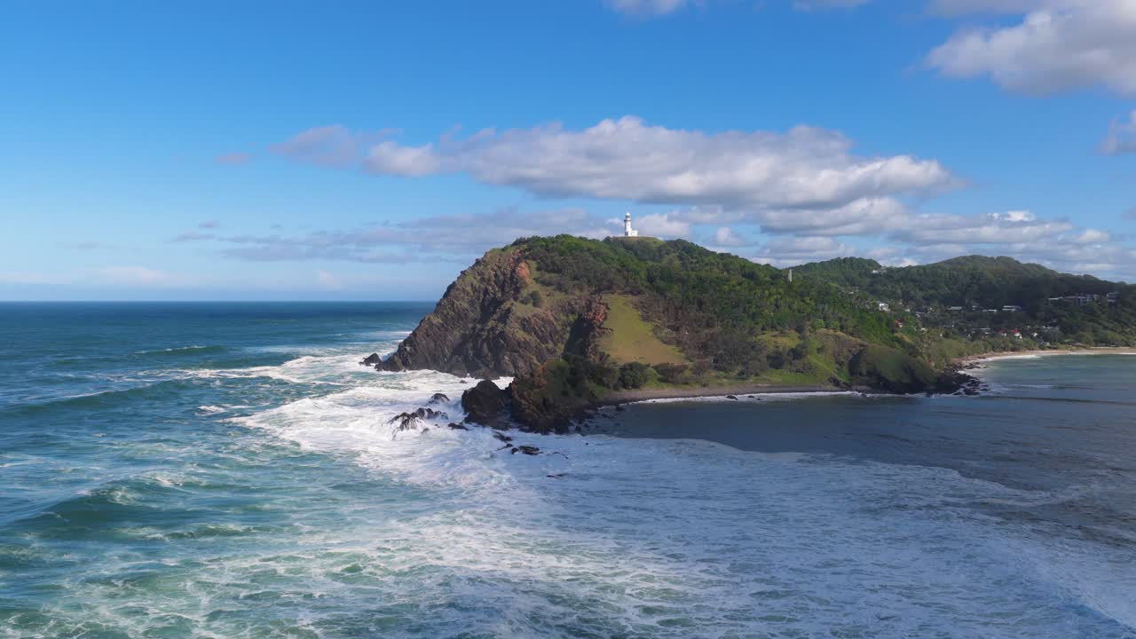 Aerial footage of Byron Bay's coastline, showcasing waves crashing against rocky cliffs under a bright blue sky