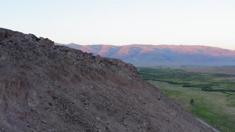 Aerial-view-of-beautiful-landscape-of-rocky-hills-and-green-fields-with-mountains-on-the-horizon-touched-by-light-from-setting-sun