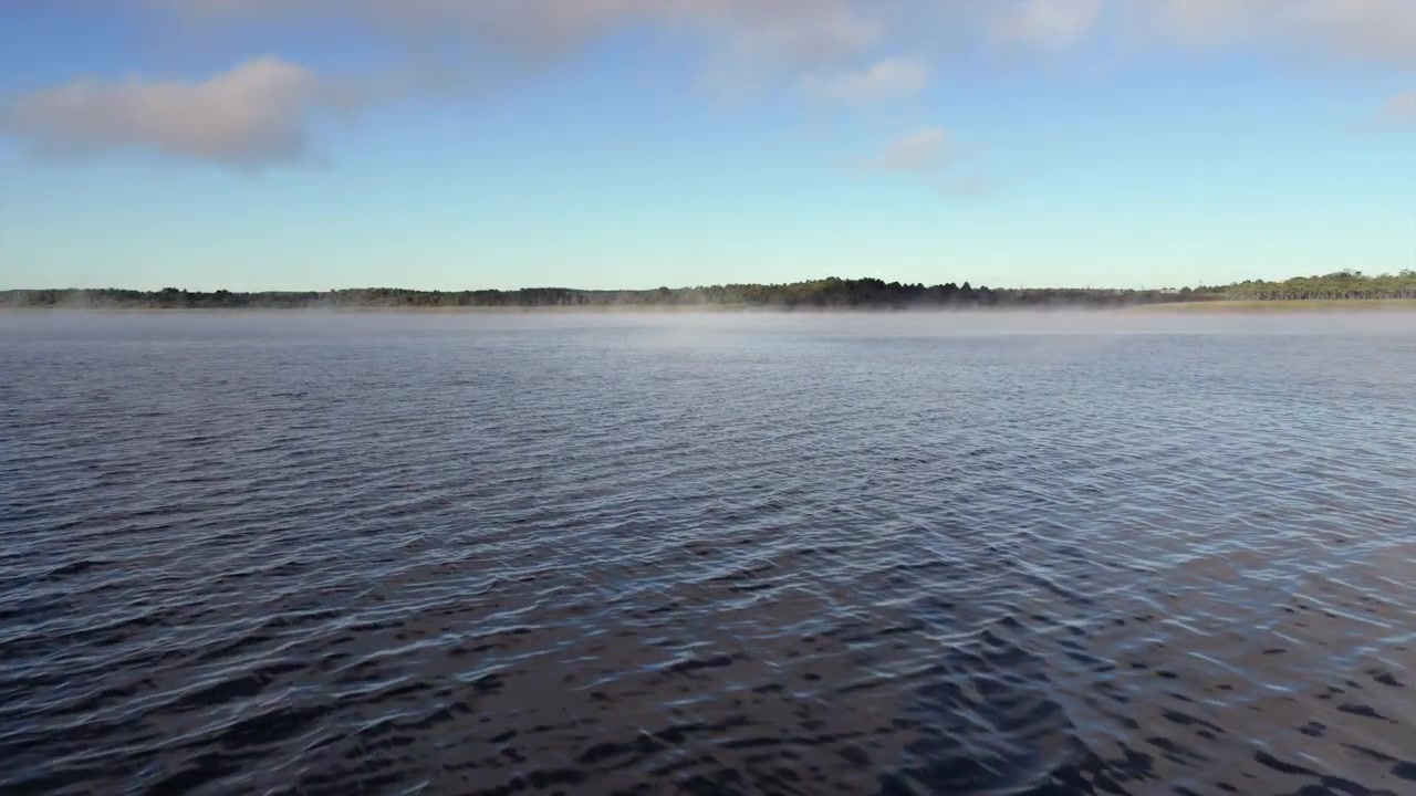 Dron vuela bajo sobre agua tranquila hacia un horizonte de bosque distante bajo nubes matutinas dispersas
