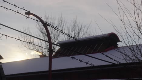 barbed wire fence in the foreground, with a snow-covered building roof and bare tree branches against a cloudy sky in the background