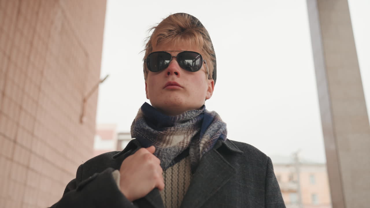 Creatore di moda maschio bianco in un passaggio ad arco, che cammina attraverso un arco con cappotto lungo e sciarpa, occhiali da sole che riflettono la città, postura sicura, rallentatore, atmosfera da campagna di moda, ritratto editoriale.