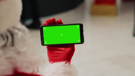 asistente en una boutique de moda decorada festiva vestido como santa claus con un teléfono inteligente con llave de croma, navegando por el sitio web de comercio electrónico de la tienda de ropa, ingresando ventas promocionales de navidad en línea