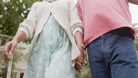 mid section of senior african american couple holding hands and walking in garden, slow motion