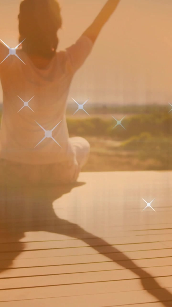 Zvedání kamery, žena se protahuje pro wellness, nohy zkřížené na dřevěné terase, kolem ní se třpytí.